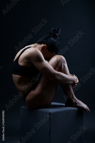 Photo of a young sports woman athletic build in a swimsuit. Theme sports and bodybuilding. Woman on a black background sits curled up