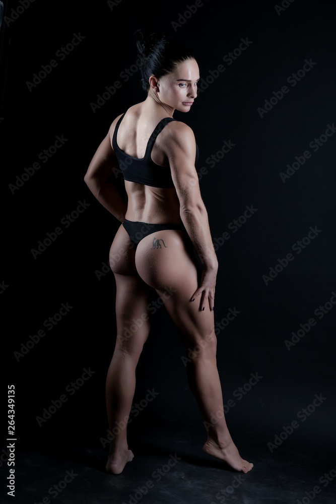 Photo of a young sports woman athletic build in a swimsuit. Theme sports and bodybuilding. Woman on a black background stands back to full growth