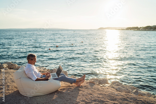 Senior man working on his laptop lying on deck chair on the beach during sunset