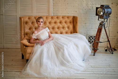 A beautiful petite blonde is sitting on the couch. The bride in a lace dress. Bride's morning