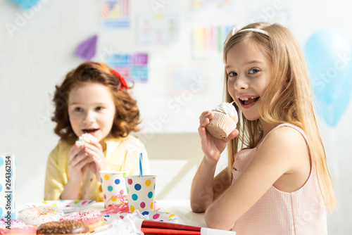 adorable kids sitting at party table and eating cupcakes during birthday celebration