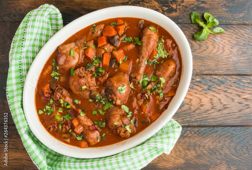 Coq au Vin, traditional French recipe of chicken braised in red wine with carrot and mushrooms. White casserole on wooden table, top view