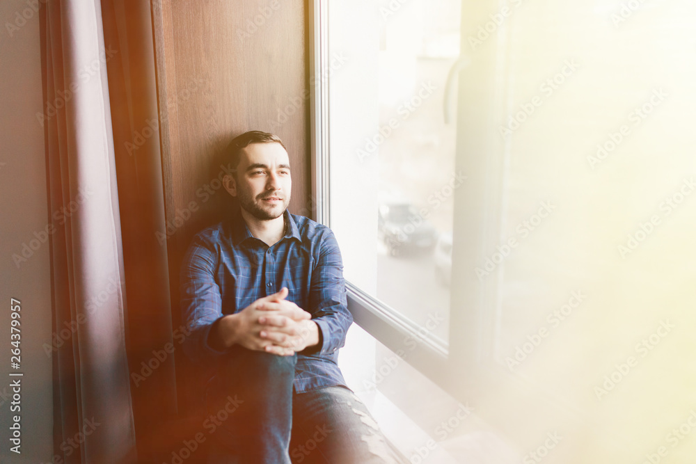 Handsome young man sitting on windowsill at home Handsome young man sitting on windowsill at home