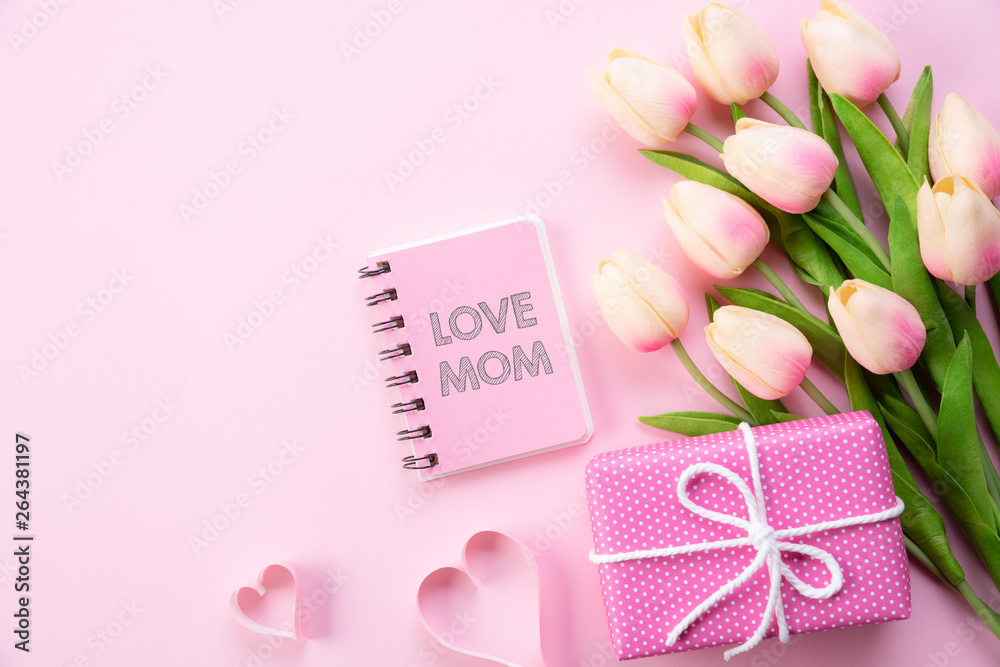 Happy mothers day concept. Top view of pink tulip flowers, gift box and note book with LOVE MOM text on pink pastel background. Flat lay.