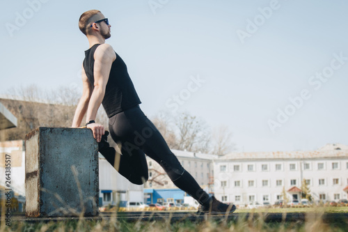A slender young man in black clothes and sunglasses is exercising outdoors. fitness athlete on the sports field. training with projectiles. warm up stretching legs. body preparation for the summer