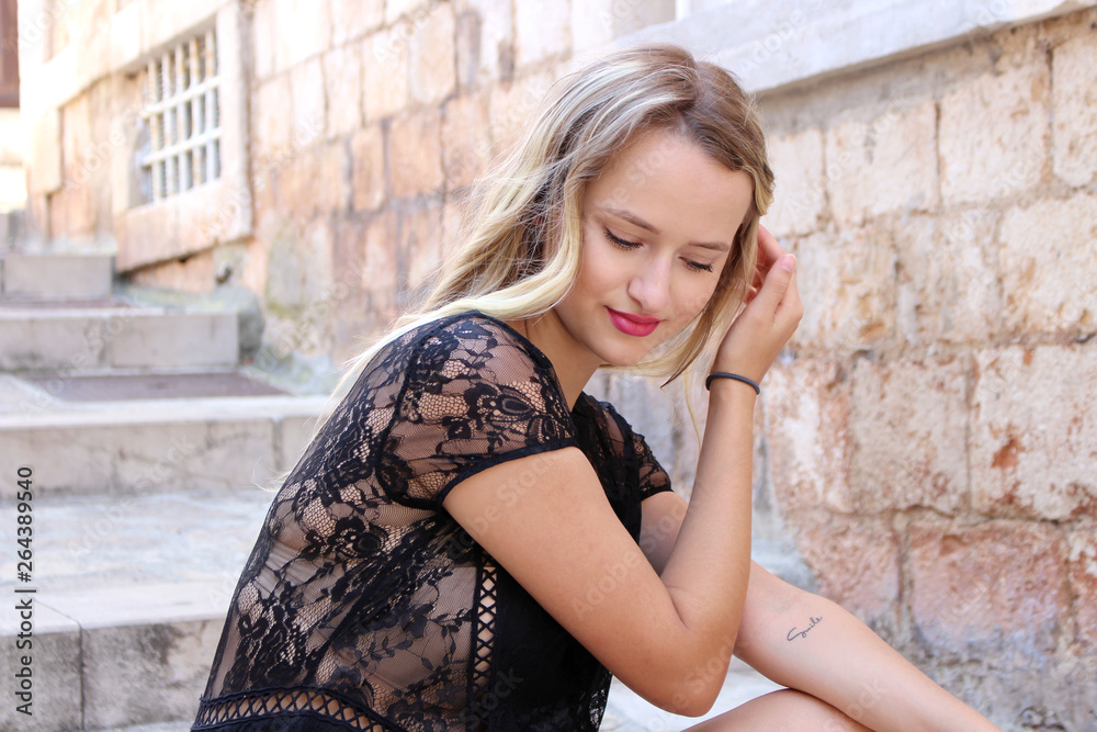 Beautiful romantic young blonde woman with blue eyes in sexy lips sits on steps in a city . She is wearing a black dress. The concept of outdoor relaxing. Beautiful romantic young blonde woman with blue eyes in sexy lips sits on steps in a city . She is wearing a black dress. The concept of outdoor relaxing.