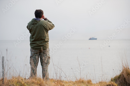 A wildlife watcher looks out to sea dressed in camouflage clothing. A boat is on the distant horizon- Image © Tony Skerl
