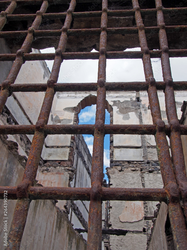 view through rusty iron bars on a window of a ruined collapsing building with crumbling walls and open to the sky view through rusty iron bars on a window of a ruined collapsing building with crumbling walls and open to the sky