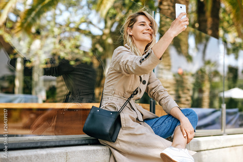 Side view of beautiful young woman smiling while holding smartphone and making self portrait, strolling outdoor in the city street against palms. Happy female taking selfie on mobile phone. Travel