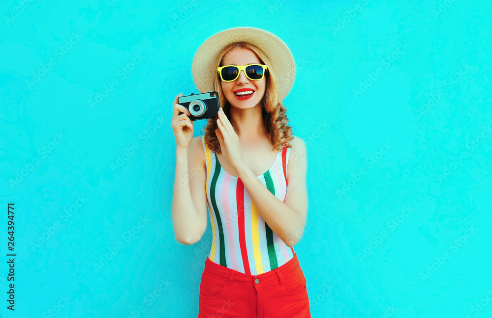 Colorful happy smiling young woman holding retro camera in summer straw hat having fun on blue wall background Colorful happy smiling young woman holding retro camera in summer straw hat having fun on blue wall background