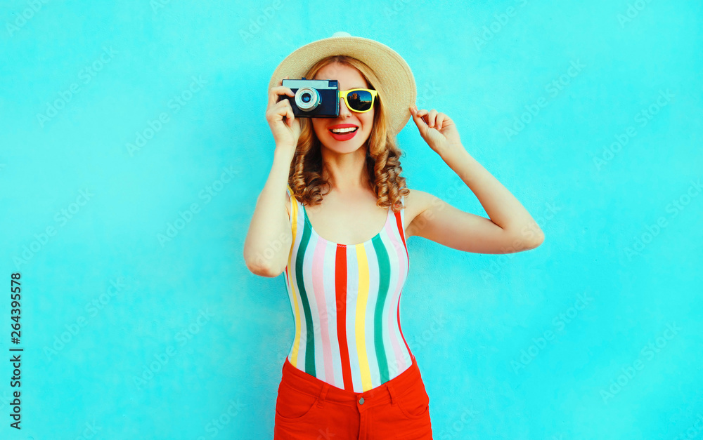 Colorful happy smiling young woman holding retro camera in summer straw hat having fun on blue wall background