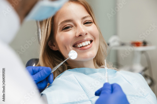 Fototapet Image of satisfied woman sitting in dental chair while professional doctor fixin