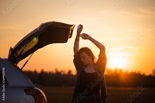 Beautiful young woman happy and dancing in a car's trunk during a road trip in Europe in the last minutes of Golden Hour sunset