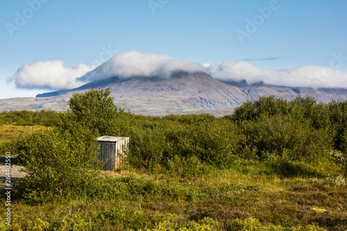 A small wooden booth standing in the middle of green grass and bushes with a mountain with a peak in the clouds in the background. Mountains, blue sky and clouds, Icelandic nature, Iceland.