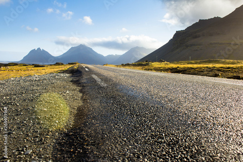 The number one ring road around Iceland in the light of the bright day sun and glare from it. Mountains and green grass on either sides of the road  the silhouettes of the mountains in the background.