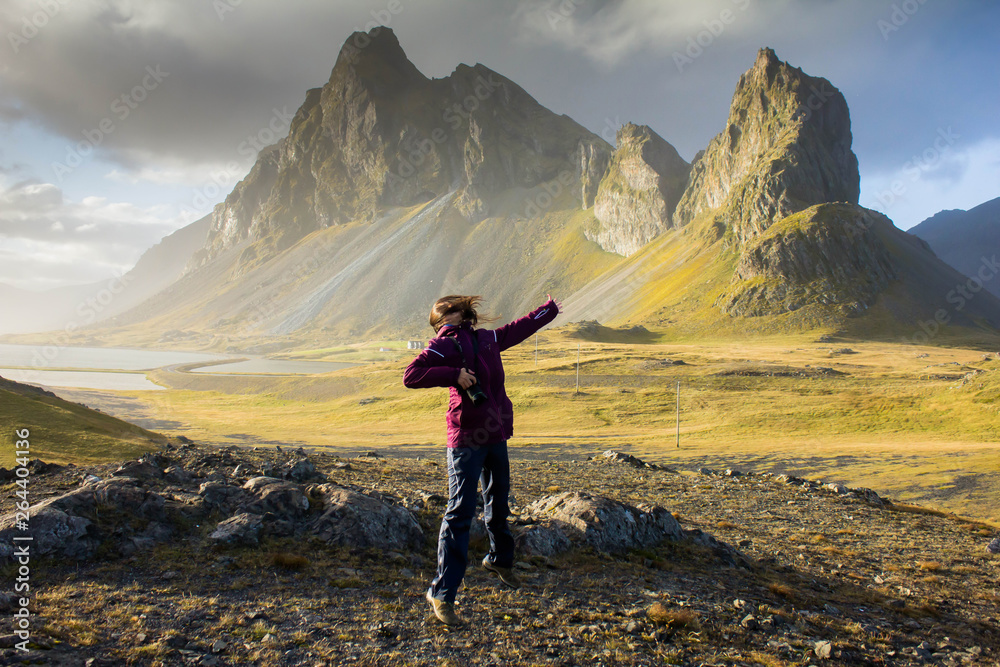 Fototapeta premium Happy joyful girl with long and dark hair jumping on nature in a outdoor hiking trip against the background of huge pointed, peaked mountains in eastern Iceland near the coast of the Atlantic Ocean.