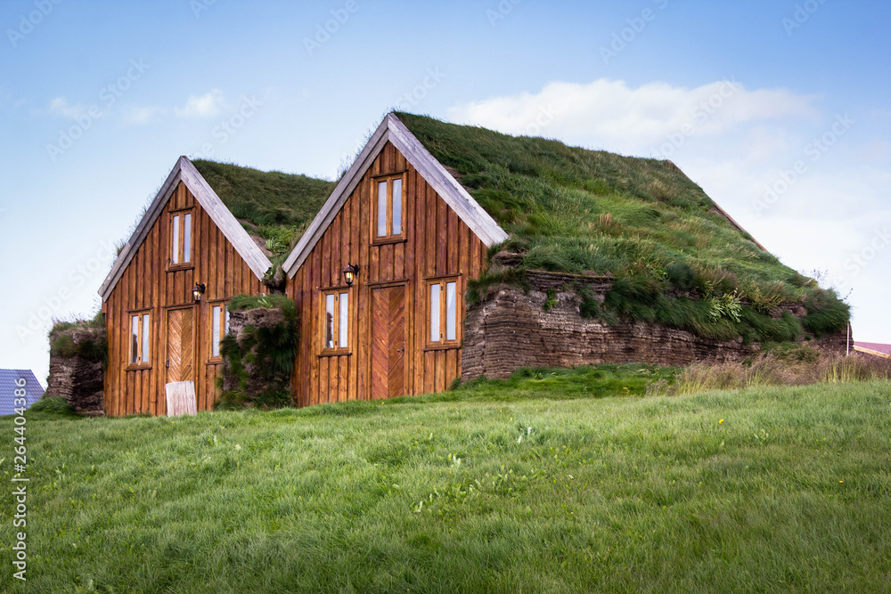 The pair of small Icelandic houses with a grass roof and brown wooden facades with three windows in the middle of the grass and blue sky above in Iceland. Countryside view and traditional architecture
