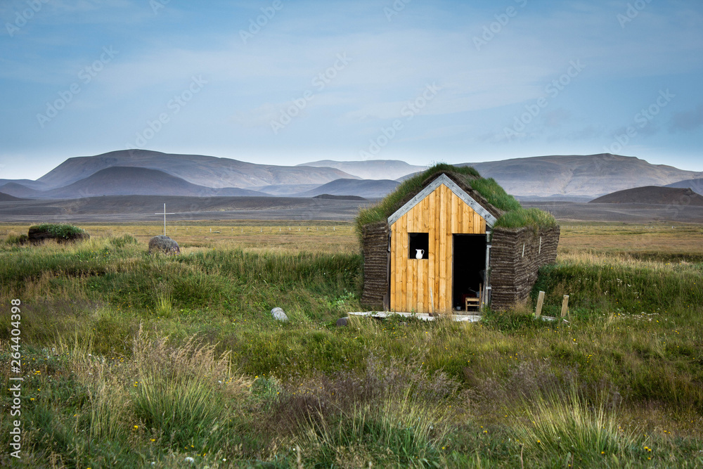 A small Icelandic house with a grass roof and a jug of milk on the window in the middle of the grass against the backdrop of foggy mountains near the grave with a cross in Iceland. Countryside view.
