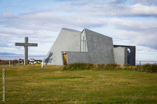 Unusual gray church in the middle of the meadow and green grass in the north of Iceland. Modern Scandinavian Icelandic church architecture. Summer trip around Iceland. Cross standing near the church.