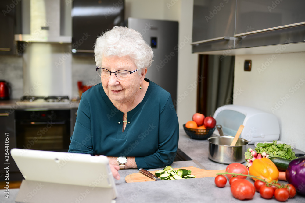 happy senior active woman cooking at home in modern kitchen and looking for recipe on a internet computer tablet happy senior active woman cooking at home in modern kitchen and looking for recipe on a internet computer tablet
