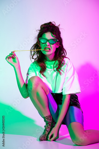 curly young woman in sunglasses sitting with bubble gum on pink with illumination