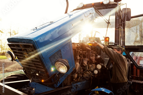 Farmer mechanic repairing blue tractor engine. Open tractor hood, engine. Repair agricultural technology at sunset.