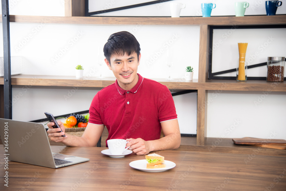 The business man with casual red t-shirt messaging on mobile phone, drinking coffee and eating sandwich, Young man working on labtop or computer in the kitchen room, loft style The business man with casual red t-shirt messaging on mobile phone, drinking coffee and eating sandwich, Young man working on labtop or computer in the kitchen room, loft style