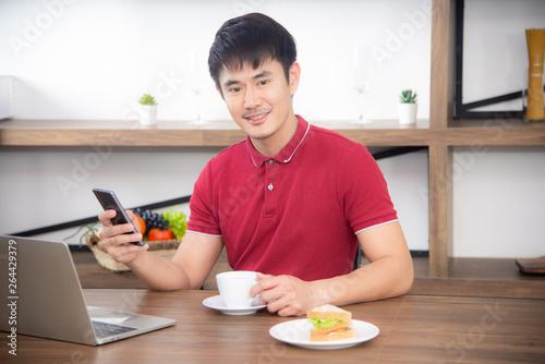 The business man with casual  red t-shirt messaging on mobile phone  drinking coffee and eating sandwich  Young man working on labtop or computer  in the kitchen room  loft style