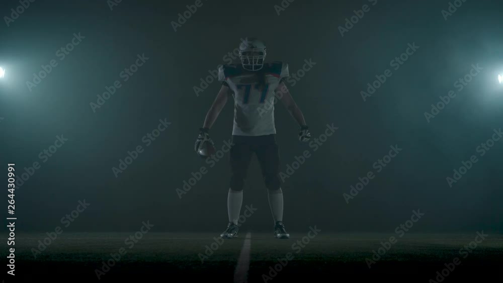 Portrait american football sportsman player in football helmet standing on the field on black background in a cloud of smoke with the ball in hands. The man raising ball makes making threatening Portrait american football sportsman player in football helmet standing on the field on black background in a cloud of smoke with the ball in hands. The man raising ball makes making threatening