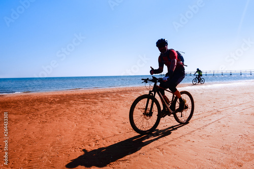 Man cycling on sport bike showing thumb up on ocean background - Silhouette of people riding bicycle on the beach shore with morning backlight - Lifestyle concept of healthy holiday activity - image