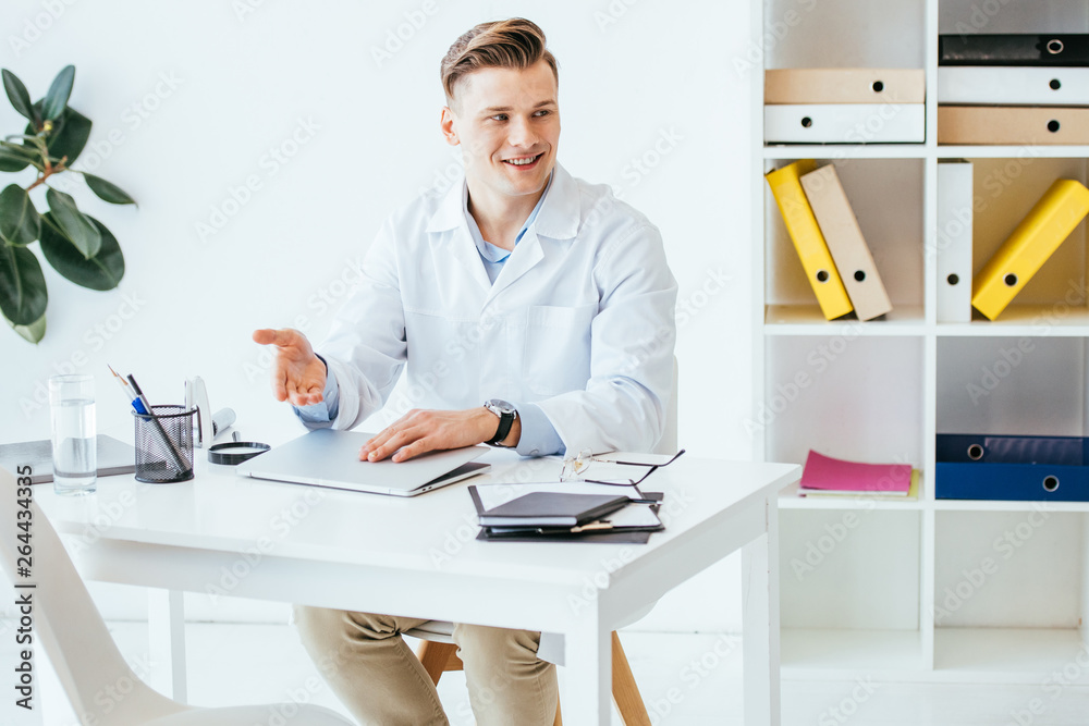 cheerful and handsome doctor in white coat gesturing near laptop in clinic