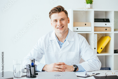 cheerful and handsome doctor in white coat sitting with clenched hands in clinic