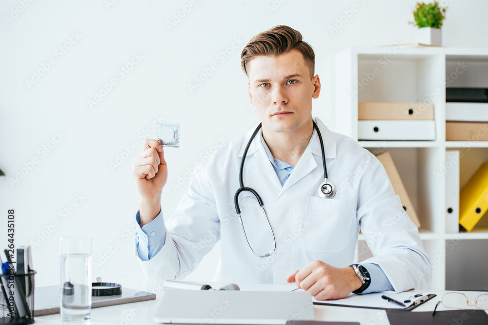 handsome and serious doctor in white coat holding condom in hand handsome and serious doctor in white coat holding condom in hand