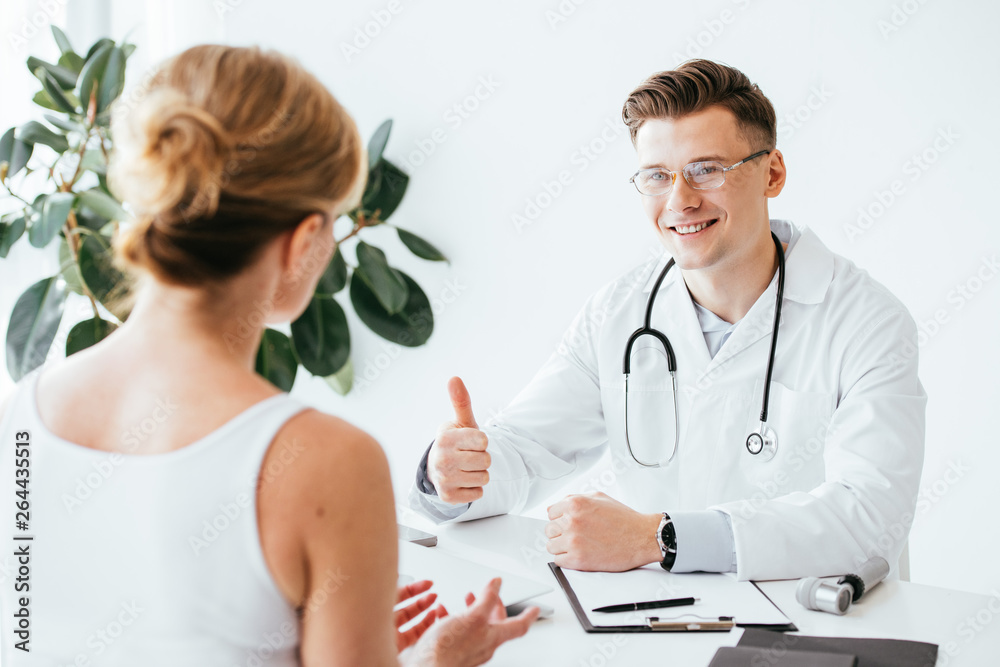 selective focus of cheerful doctor in glasses showing thumb up while looking at patient selective focus of cheerful doctor in glasses showing thumb up while looking at patient