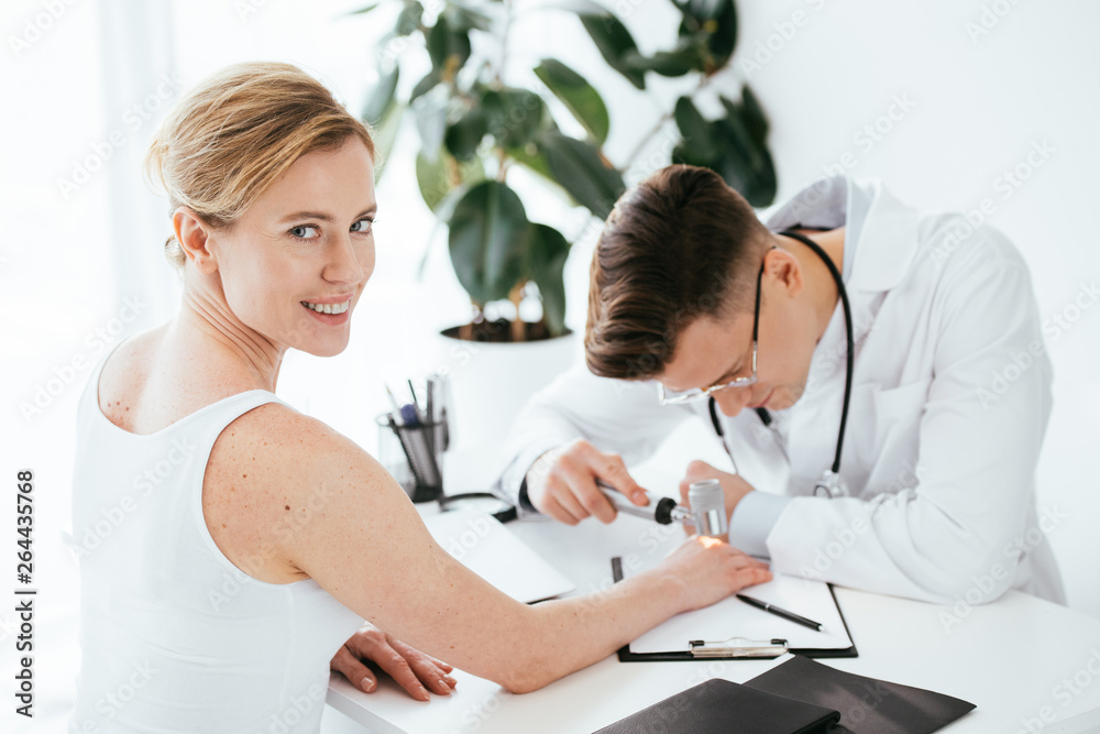 Obraz premium selective focus of cheerful woman looking at camera while dermatologist holding dermatoscope