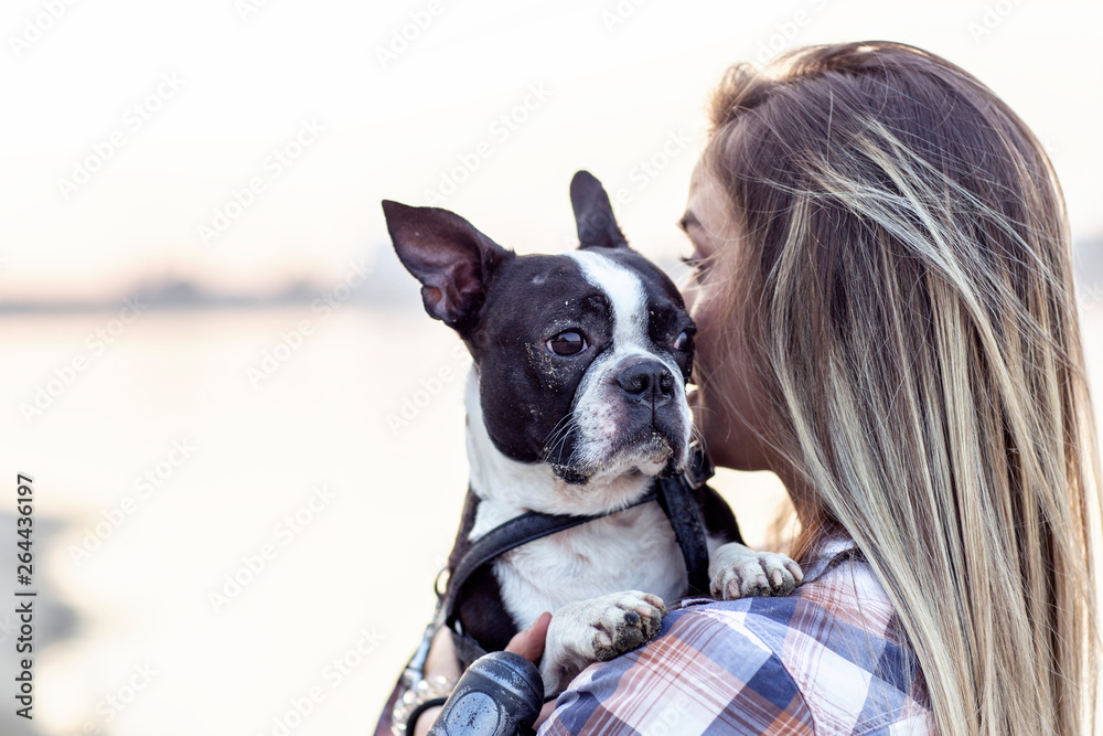 Beautiful girl holding on her hands boston terrier black and white dog on sandy beach near the river