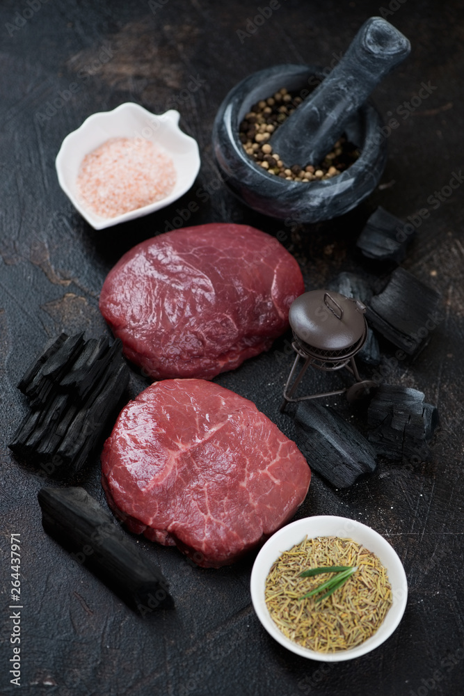 Raw marbled beefsteaks with condiments and charcoal over dark brown stone background, vertical shot Raw marbled beefsteaks with condiments and charcoal over dark brown stone background, vertical shot