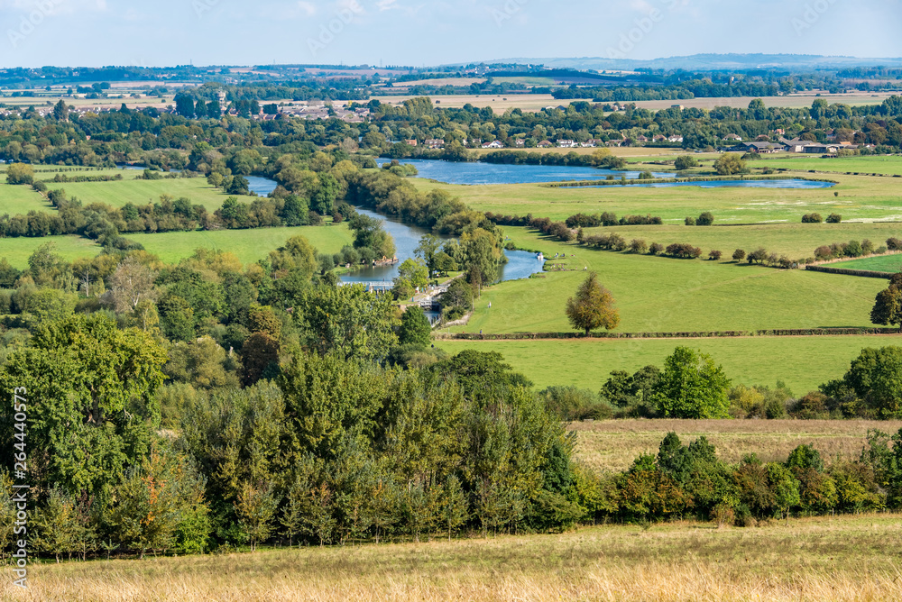 A sunlit rural scene with green fields and a river, seen from above. It is the River Thames seen from the hills known as the Wittenham Clumps, in Oxfordshire, United Kingdom. 