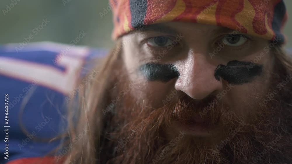 Close up portrait of a bearded american football player with long hair and beard in sports equipment puts black paint on his face. The man expressing his aggression. Front view