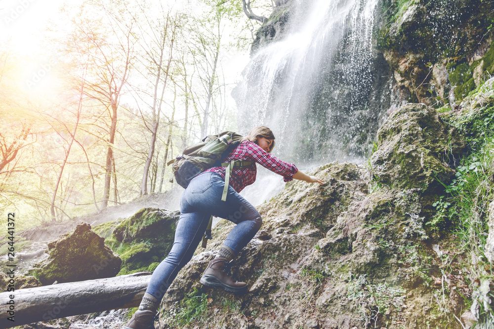 young woman with backpack hiking near waterfall in forest 
