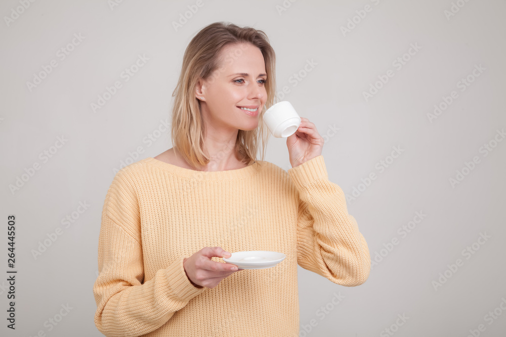 Beautiful young tender girl with blond hair drinks delicious coffee she is dressed in a yellow sweater. poses against a gray background. facial expression, emotion. Beautiful young tender girl with blond hair drinks delicious coffee she is dressed in a yellow sweater. poses against a gray background. facial expression, emotion.