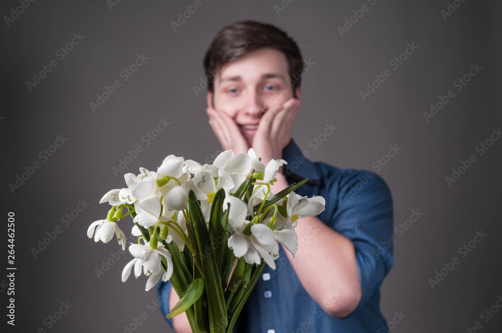 selective focus of handsome surprised and smiling young man in blue shirt touching cheeks and looking at bouquet of snowdrops on grey background selective focus of handsome surprised and smiling young man in blue shirt touching cheeks and looking at bouquet of snowdrops on grey background