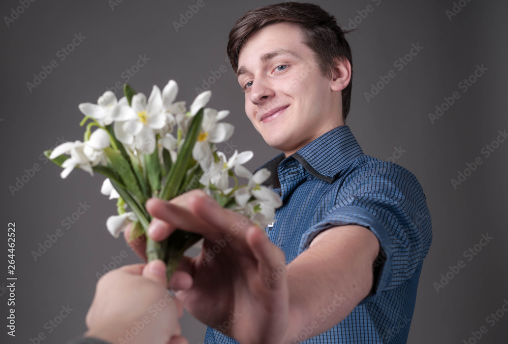 selective focus of handsome surprised and smiling young man in blue shirt taking bouquet of snowdrops from woman hand on grey background selective focus of handsome surprised and smiling young man in blue shirt taking bouquet of snowdrops from woman hand on grey background