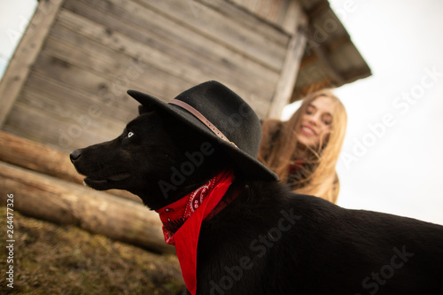 Happy young woman plaing with her black dog in fron of old wooden house. Girl tries a hat to her dog photo