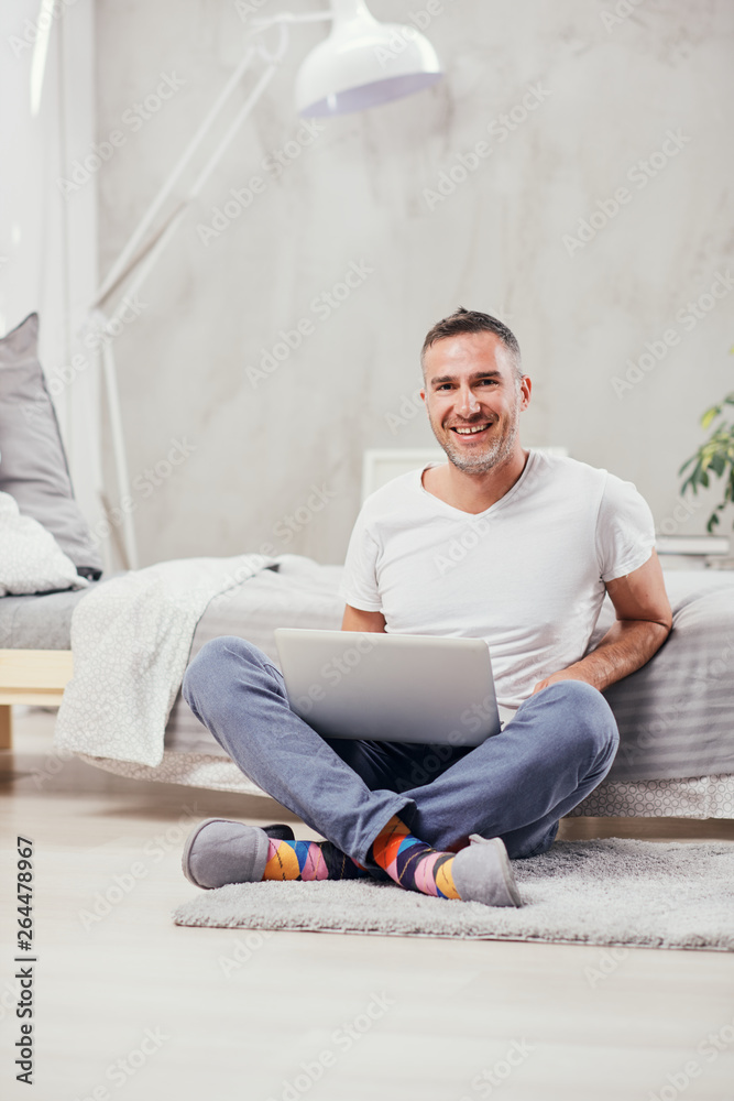 Handsome middle aged man sitting on the floor wirh crossed legs, leaning on bed and using laptop.