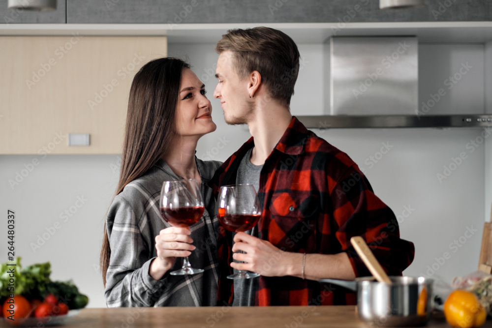 Beautiful young couple in plaid shirts in the kitchen preparing food and drinking red wine from glasses. Beautiful young couple in plaid shirts in the kitchen preparing food and drinking red wine from glasses.