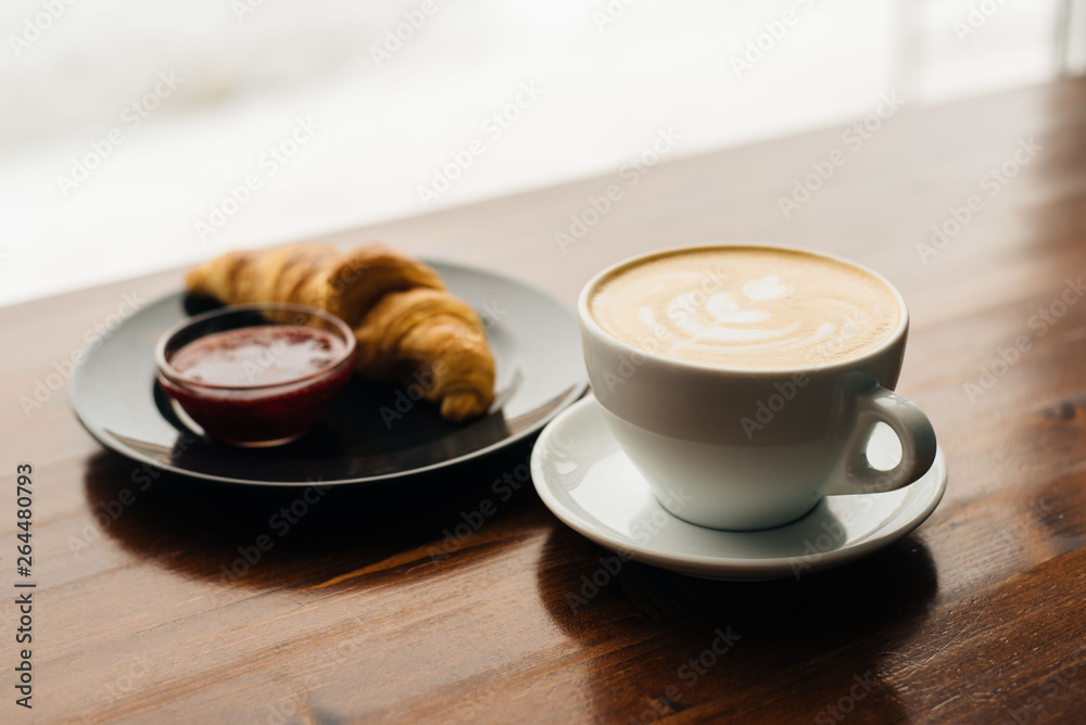 Fototapeta Cup of cappuccino with latte art and croissant with jam on the wooden table in cafe in the morning