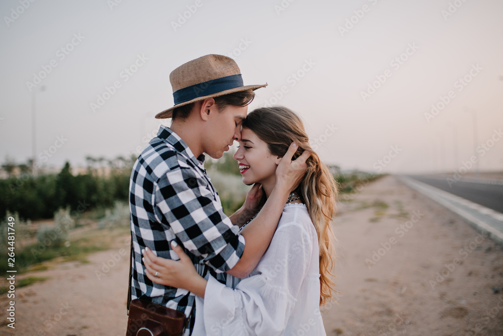 Excited cheerful girl in white tunic soft embracing boyfriend in stylish shirt, while he touching her long hair. Young couple of travelers hugging standing near the road and enjoys evening date