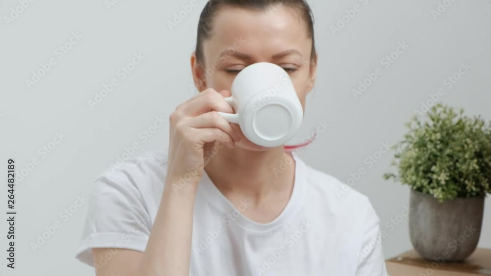 Young beautiful woman in jeans and a white T-shirt is drinking coffee while sitting on the floor against a background of cardboard boxes. The concept of relaxation on the day of the move. Young beautiful woman in jeans and a white T-shirt is drinking coffee while sitting on the floor against a background of cardboard boxes. The concept of relaxation on the day of the move.