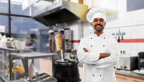cooking, profession and people concept - happy male indian chef in toque with crossed arms over kebab shop kitchen background